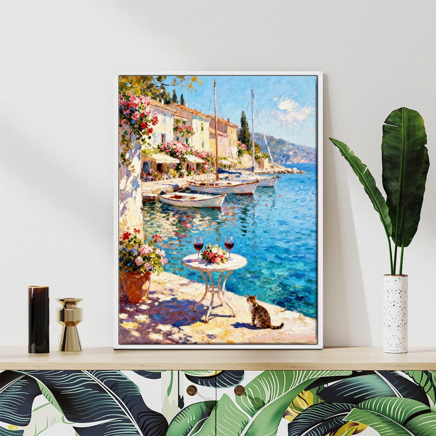 white frame wall art canvas painting showing a sunny seaside town with a table of red wine and flowers on the pier and a cat.